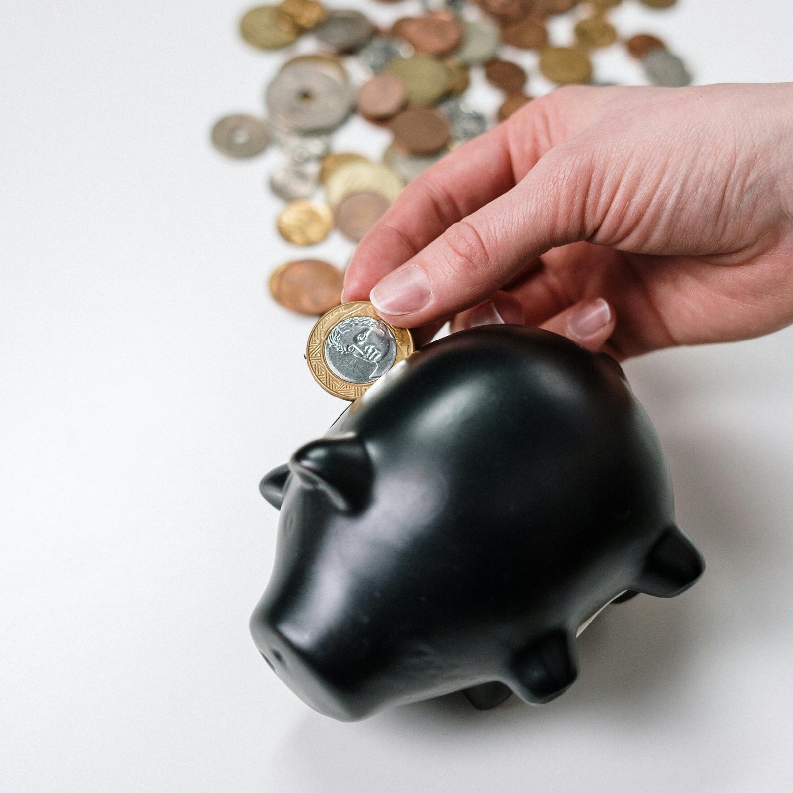 A hand putting a coin into a black piggy bank with scattered coins on a white background.